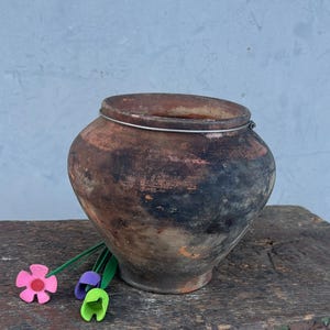 May include: A weathered, brown and black ceramic pot with a lid, placed on a rustic wooden bench. The bench is painted dark brown with visible wood grain. Small felt flowers in pink, purple, and green provide a splash of colour.