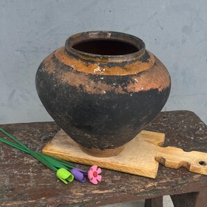 May include: A rustic, dark-colored ceramic pot with orange and black bands sits on a small, weathered wooden table. A wooden cutting board and colorful paper flowers are also on the table. The background is a muted gray.