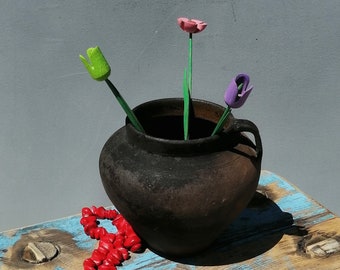 May include: A rustic wooden stool with blue paint and a red beaded necklace. A dark brown ceramic pot with three wooden flowers in pink, green, and purple sits on top of the stool.