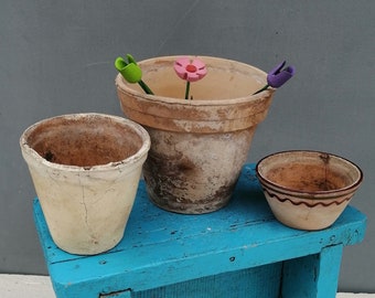 May include: Three terracotta pots with colorful flower accents sit on a blue wooden stool. The stool is on a white wooden surface.