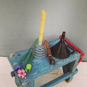 Puede incluir: Un taburete de madera azul con un batidor de metal, un pequeño embudo, un collar de cuentas rojas y tres flores de plástico de colores.