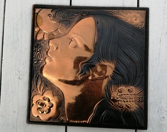 May include: A copper wall plaque with a relief sculpture of a woman's face. The woman has long dark hair and is wearing a flower behind her ear. The plaque is set against a white wooden background.
