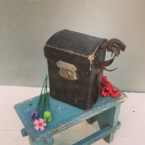 May include: A small, dark brown wooden box with a metal latch and a leather strap sits on a blue painted wooden stool. The stool is on a white wooden surface. There are three small plastic flowers in green, purple, and pink on the stool.