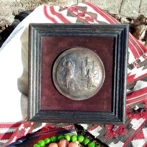 May include: A framed, circular, bronze-colored plaque with a raised image of two figures and a building. The plaque is set against a dark red velvet background. The frame is dark brown wood.