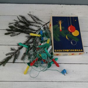 May include: A box of vintage Christmas lights with a blue and yellow design. The box says "Elektrogiрлянда" and features a stylised Christmas tree with a star, a red ball, and a yellow ball. The lights are scattered around the box, including green, red, yellow, and white bulbs.