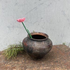 May include: A rustic, dark brown ceramic pot with a pink flower and green stem. The pot sits on a weathered, brown wooden bench. The background is a light gray wall.