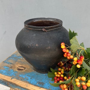 Peut inclure: Un pot en terre cuite noir rustique posé sur un tabouret en bois bleu et brun. Une branche de baies rouges et jaunes avec des feuilles vertes repose sur le tabouret à côté du pot.