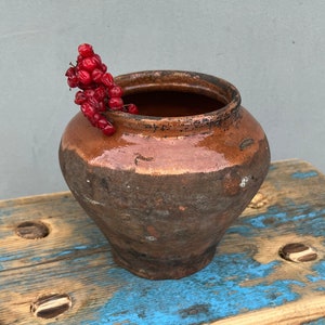 Könnte beinhalten: Ein rustikaler brauner Tontopf mit einer breiten Öffnung, der auf einem blauen Holzhocker steht. Ein Zweig mit roten Beeren liegt im Topf.