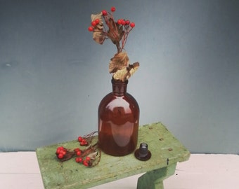 May include: A brown glass bottle with a red berry sprig inside sits on a green wooden stool. The stool has a rustic, distressed look.