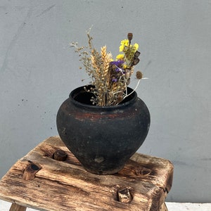 May include: A rustic wooden stool with three legs, featuring a dark brown ceramic pot with dried flowers and wheat stalks.