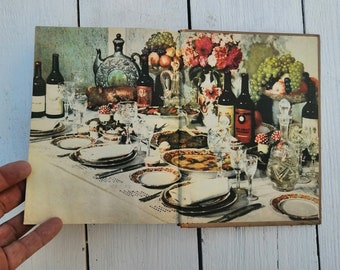 Puede incluir: Un libro de cocina vintage abierto en una página que muestra una mesa puesta con una variedad de platos, botellas de vino y vasos. La mesa está cubierta con un mantel blanco con un patrón de encaje. Hay flores y frutas en el fondo.