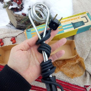 Puede incluir: Un calentador de inmersión eléctrico plateado con un cable y un enchufe negros. El calentador está en espiral y tiene un mango de plástico negro. El calentador está en su caja original, que es amarilla y azul con el texto "Электрипо" en cirílico.
