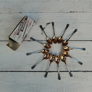 Puede incluir: Un juego de 12 cucharaditas doradas dispuestas en círculo sobre una superficie de madera blanca. Las cucharas tienen mangos negros y están etiquetadas "Столовые Приборы" que se traduce como "Vajilla" en español.