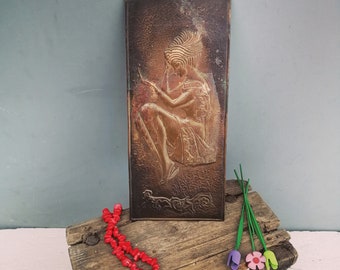May include: A rectangular copper relief sculpture depicting a seated figure with ornate headdress and clothing. The artwork is displayed on a rustic wooden surface, accompanied by a red beaded necklace and colorful felt flowers.