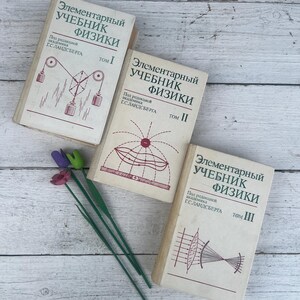 Op de afbeelding: Drie vintage natuurkundeboeken met Russische tekst, getiteld "Elementair Leerboek Natuurkunde", met illustraties op de covers. De boeken zijn gerangschikt met een paar decoratieve bloemen op een licht houtnerfoppervlak.