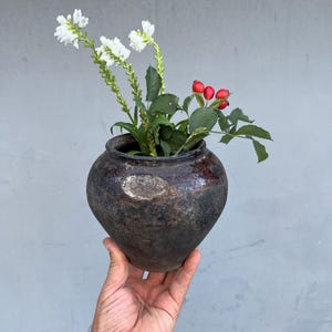 Puede incluir: Una olla de cerámica antigua de color marrón oscuro llena de flores blancas y bayas rojas. La olla tiene forma redondeada y un aspecto desgastado, sostenida por una mano sobre un fondo gris. Las flores y las bayas añaden un toque de color a la estética rústica.