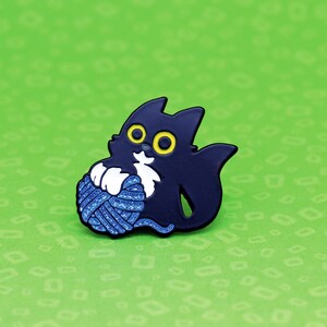 May include: Black enamel pin of a cat playing with a blue ball of yarn. The cat has white fur on its chest and yellow eyes.