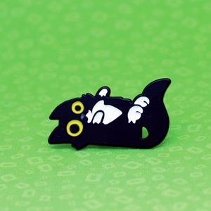 May include: Black enamel pin of a cartoon cat with yellow eyes, lying on its back with its paws in the air.