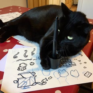 Op de afbeelding: Een zwarte kat rust op een oppervlak met een wit vel papier met zwarte inktillustraties van een teddybeer, een geschenkdoos, ballonnen en diverse vormen, met een zwarte digitale stylus rechtop in een cilindrische houder.