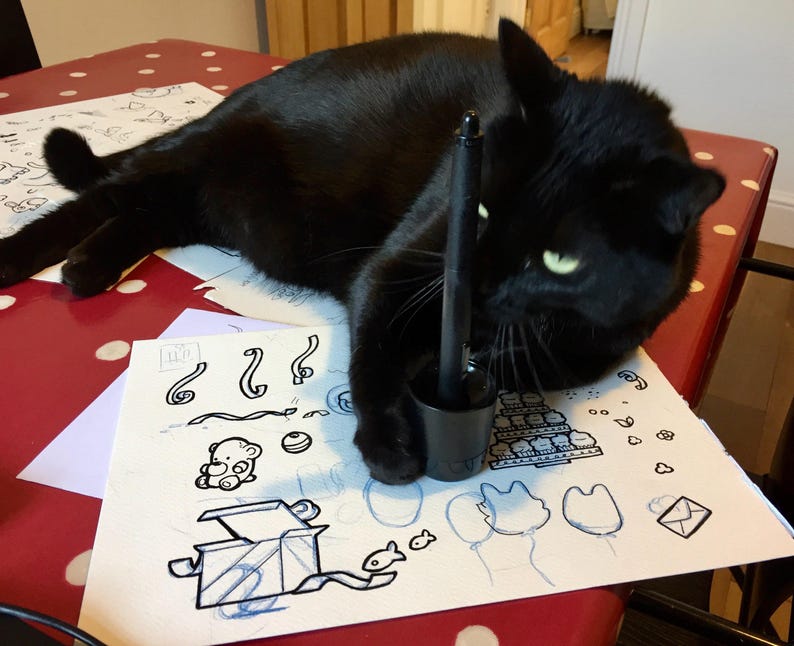 Op de afbeelding: Een zwarte kat rust op een oppervlak met een wit vel papier met zwarte inktillustraties van een teddybeer, een geschenkdoos, ballonnen en diverse vormen, met een zwarte digitale stylus rechtop in een cilindrische houder.