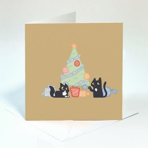 May include: A Christmas card featuring two black cats with white markings, one holding a blue bauble and the other sitting next to a wrapped gift, in front of a green Christmas tree with silver tinsel and colourful baubles.