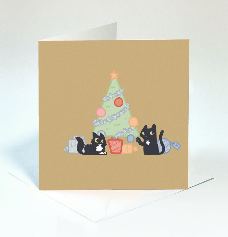 May include: A Christmas card featuring two black cats with white markings, one holding a blue bauble and the other sitting next to a wrapped gift, in front of a green Christmas tree with silver tinsel and colourful baubles.