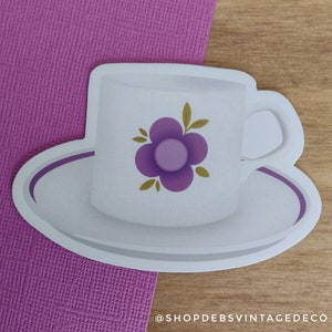 May include: A white sticker of a teacup and saucer with a purple floral design. The teacup has a handle and sits on a matching saucer. The sticker is on a wooden surface with a purple textured background.