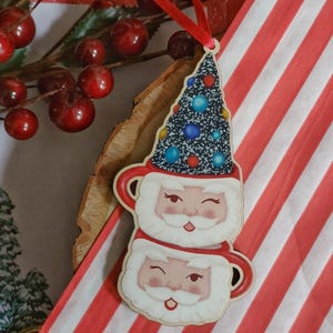 May include: A wooden Christmas ornament shaped like two stacked mugs with Santa faces. The top mug has a Christmas tree hat with colourful ornaments.