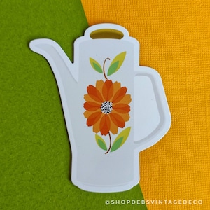 May include: White ceramic pitcher with a floral design. The pitcher has a large orange flower with a black center and green leaves. The pitcher has a spout and a handle.