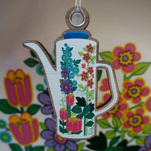 May include: A wooden keychain shaped like a teapot with a floral design. The teapot is white with blue, green, pink, and yellow flowers. The keychain has a clear plastic ring.
