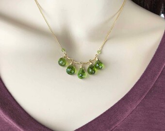 May include: A gold chain necklace with a delicate design features five teardrop-shaped green gemstones hanging from the chain.