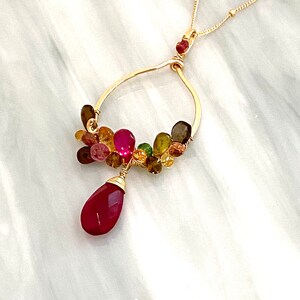 May include: A gold necklace with a teardrop-shaped pendant made of red gemstone. The pendant is surrounded by a cluster of smaller gemstones in various colors, including pink, green, brown, and yellow.