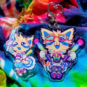 May include: Two colorful acrylic keychains featuring cartoon cat characters with glasses and accessories. The keychains are on a tie-dye fabric inside a pink container, with a star and heart-shaped keychain clasp.