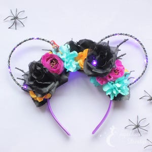 Puede incluir: Diadema con luces moradas con flores negras, rosas, amarillas y turquesas. La diadema está decorada con ramas de alambre negro.