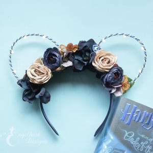 Könnte beinhalten: Leuchtende Mäuseohren-Haarreifen mit einem floralen Design aus blauen, beigen und braunen Blumen. Der Haarreifen ist mit einer blauen und weißen Lichterkette verziert.