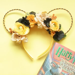 May include: A yellow headband with floral accents, including yellow, black, and cream-colored flowers. The headband has a gold-colored base and a decorative twisted wire design. A book with the title "Harry Potter and the Prisoner of Azkaban" is visible.