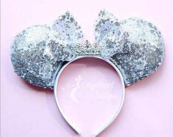 May include: Silver sequin Minnie Mouse ears with a silver crown headband. The ears are shaped like Minnie Mouse's ears and have a bow on top. The headband is decorated with a silver crown.
