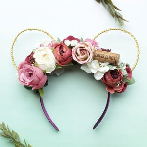 Op de afbeelding: Een bloemenhoofdband met gouden draadaccenten in de vorm van oren. De hoofdband heeft een assortiment roze, bordeauxrode en witte bloemen, met een kurk met "LOUIS JADOT". De hoofdband heeft een paarse basis.