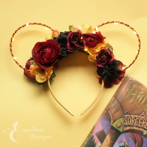 May include: A floral headband with glowing lights, featuring burgundy and yellow flowers. The headband has gold-colored wire and a book with the title "Harry and the Sorcerer" is visible in the background.