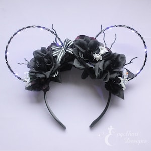 May include: Black and white floral headband with light-up wire ears. The headband is decorated with black roses, white flowers, and a black and white striped bow.