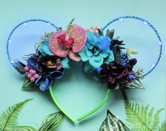 Peut inclure: Oreilles de Minnie Mouse LED bleu clair avec un bandeau floral composé de fleurs roses, bleues et violettes. Le bandeau est décoré de différentes fleurs, notamment des orchidées, des hortensias et des roses.