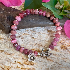 May include: A pink gemstone bracelet with a silver flower charm and a silver clasp. The bracelet is made of faceted beads and is photographed on a wooden surface with pink flowers.