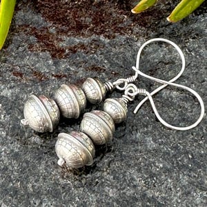 May include: A pair of silver earrings with a textured, beaded design. Each earring features four round beads with a textured surface, connected by a silver wire. The earrings are hanging from silver hooks.