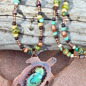 May include: A handcrafted necklace featuring a copper turtle pendant with a turquoise stone center. The necklace has a beaded chain with turquoise, brown, and green beads. The pendant is approximately 2 inches wide.