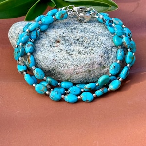 May include: A turquoise beaded bracelet with a silver clasp. The bracelet is made of oval-shaped turquoise beads strung together with silver-colored spacers. The bracelet is wrapped around a gray stone.