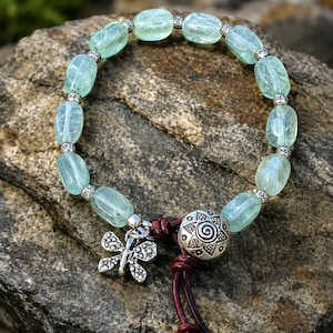 May include: A beaded bracelet featuring light blue, oval-shaped stones and silver-colored accents. A silver dragonfly charm and a decorative silver bead with a spiral design are also included. The bracelet is displayed on a textured gray rock.