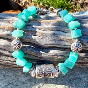 May include: A turquoise gemstone bracelet with silver accents. The bracelet features irregular-shaped turquoise beads, interspersed with silver beads and a rectangular silver charm with an elephant design. The bracelet is secured with a silver clasp.