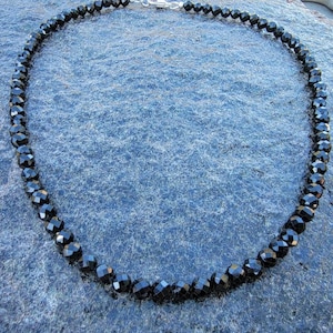 May include: A black beaded necklace made of faceted black onyx beads. The necklace is on a blue background.