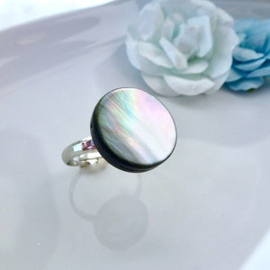 Könnte beinhalten: Ein silberner Ring mit einem großen, schillernden, runden Perlmuttstein. Der Stein zeigt eine Reihe von Farben, darunter Rosa, Grün und Blau. Der Ring ist vor einem weißen Hintergrund mit weichen floralen Akzenten platziert.