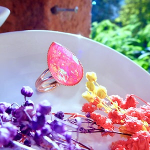 May include: A pink iridescent ring with a teardrop-shaped stone set in a gold band. The ring is displayed on a white plate with dried flowers.